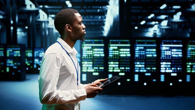 Electrician inspecting server racks delivering web content to customers over the internet. African american wireman using tablet to take note of critical systems, looking for potential errors