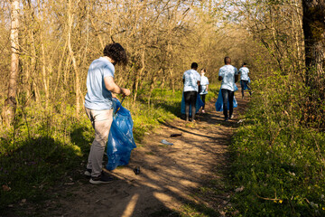 Male environmentalist collecting trash with a long claw tool and bag, grabbing plastic waste and rubbish to tidy up the forest area. Young man cleaning the natural habitat, global pollution.