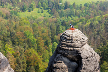 Naklejka premium Tired rock climber resting break on peak summit rocky sansdtone mountain in Saxon Switzerland National Park Germany with green forest background. Extreme endurance sports adventure outdoors activity
