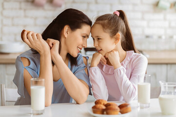 A mother and her young daughter are seated at a kitchen table, sharing a sweet moment together during breakfast. Mother is smiling and holding a pastry, while the daughter rests her chin on her hands