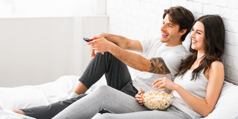 A man and woman are relaxing on a bed, watching television. They are both smiling and laughing, enjoying each others company. The man is holding a remote control and the woman is eating popcorn