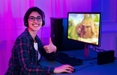 A woman in a plaid shirt sits at a computer desk with a gaming monitor displaying an animated scene. She wears headphones and smiles at the camera, giving a thumbs up