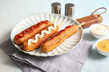 Plate with tasty corn dogs and bowl of sauces on light blue background