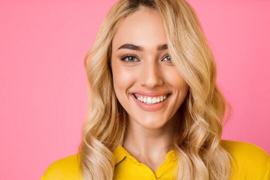 The image features a close-up portrait of a young woman with blonde hair, smiling brightly at the camera. She is wearing a yellow shirt and is positioned against a solid pink background.