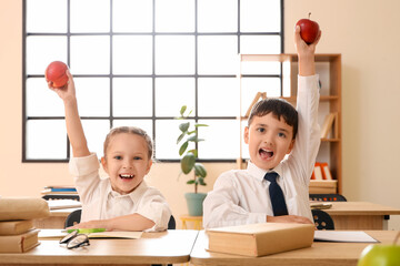 Happy little schoolchildren with apples at desks in classroom