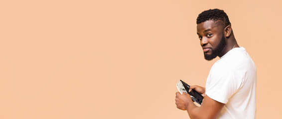 A young African American man wearing a white t-shirt stands against a beige background. He holds a wallet in his right hand, showing a glimpse of cash inside. He looks over his shoulder