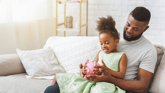 African American father and daughter are sitting on a couch together. The daughter is holding a piggy bank in her lap while the father smiles at her, copy space