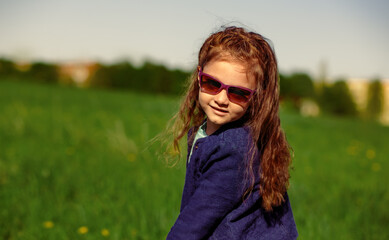 Enjoying kid girl with beautiful long curly hairstyle walking on summer blue sky and green grass outdoors background in fashion violet sunglasses. Lifestyle