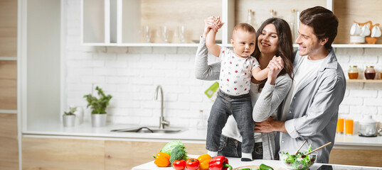 A happy family is playing in their modern kitchen while preparing food. The mother is holding up their young child, who is smiling and reaching out. Father is standing next to them, looking lovingly - Powered by Adobe