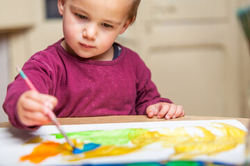 Close-up of a toddler painting with paints and a brush on a paper on a desk.