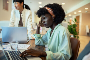 Stressed African American woman working on laptop in busy office environment
