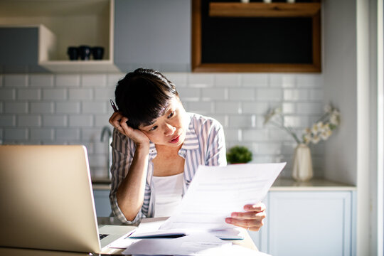 Asian woman reading bills in the kitchen with laptop at home