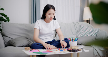 Portrait of Young Asian woman sitting on sofa decorating a greeting card in the living room, Holiday hobbies, crafts, DIY cards