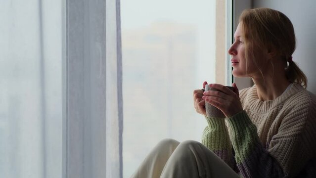 Dreamy adult woman drinks morning tea or coffee, sitting on the windowsill in a cozy apartment and draws with her finger on the window pane.