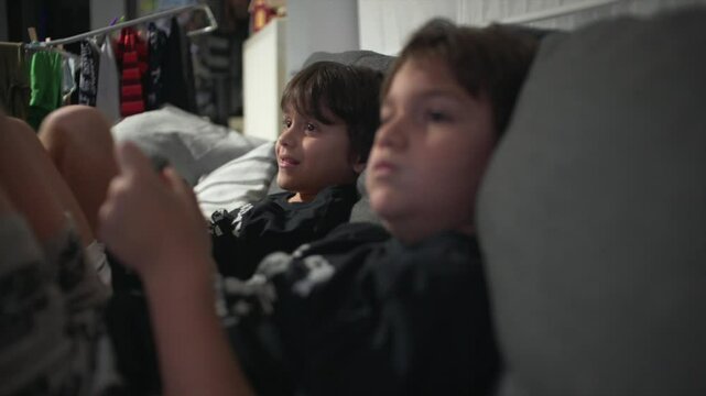 Side view close up of two caucasian boys, relaxing at home on the couch, playing video games and staring at the screen, while holding the game controllers 4K