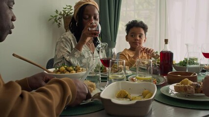 Medium shot of young African American woman with braids, in dress and 6-year-old son sitting at table at family party on holiday, enjoying festive meal, chewing on roast chicken and drinking wine