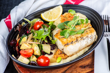A serving of grilled Barramundi, served with mashed potato, green salad in a dark oval bowl