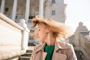 A woman with long hair is standing on a set of stairs. She is wearing a green shirt and a tan coat.