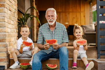 Portrait of happy grandchildren sitting on a porch with granddad and eating watermelon.