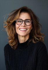 Fototapeta premium Studio portrait of a mid-40s woman with shoulder-length brown hair, laughing and wearing glasses, on a gradient dark grey background, showcasing timeless beauty and effortless chic style