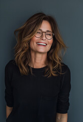 Obraz premium Studio portrait of a mid-40s woman with shoulder-length brown hair, laughing and wearing glasses, on a gradient dark grey background, showcasing timeless beauty and effortless chic style
