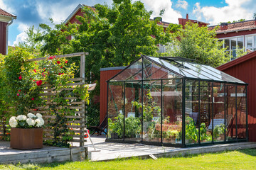 Beautiful greenhouse glass house in the garden yard near the villa. Wicker rattan chairs inside. Lots of pots with different plants.  Greenhouse for growing plant seedlings. Landscape garden design.