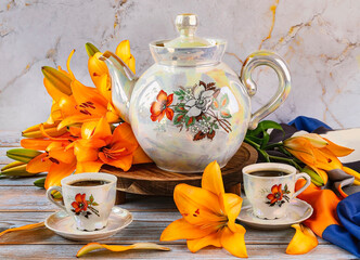 A vintage teapot with wildflowers, two white cups of coffee, a beautiful bouquet of orange lilies on the table. Summer still life with yellow flowers