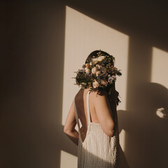 Woman in linen dress holding flowers covering her face. Shadowplay in front of beige wall....