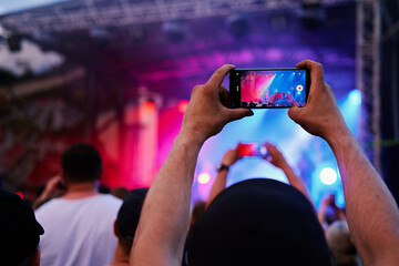 Person standing in front of stage, recording live music concert on smartphone. People crowd near stage enjoys musician band's performance