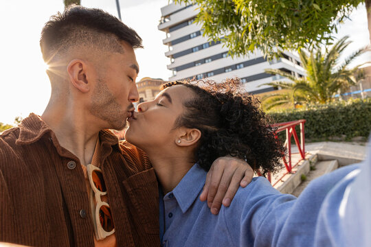 Multiracial couple kissing and taking selfie at park - Powered by Adobe