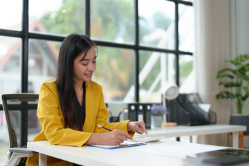 .Cheerful Asian businesswoman using laptop and writing on charger to take analytical notes. and happily collects thoughts and ideas in her office.