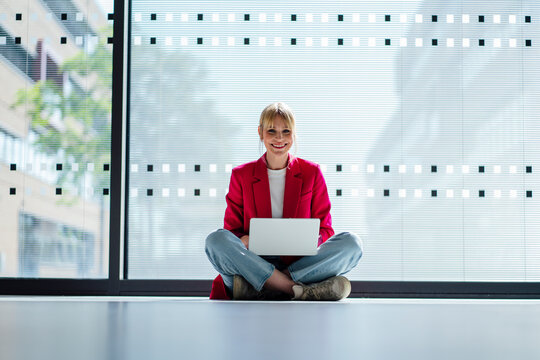 Smiling blond businesswoman sitting cross-legged with laptop on floor in front of window at office