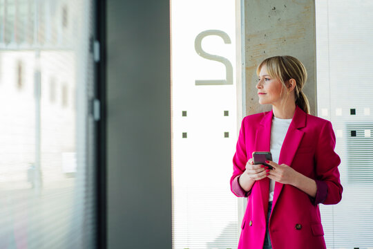 Blond businesswoman wearing pink blazer standing with smart phone looking out through office window