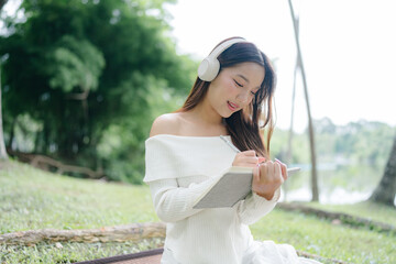Naklejka premium Side view of a cute teenager wearing headphones sitting on the grass and writing a notebook while relaxing on the weekend