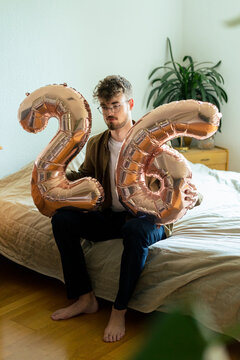Depressed young man with number 26 balloons sitting on bed at home