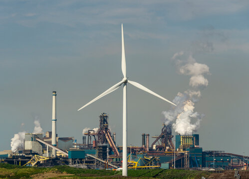 Blast furnaces emitting smoke from chimneys with spinning wind turbines under sky
