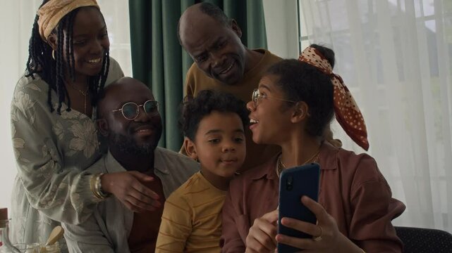 Medium close-up of cheerful Black family posing for fun selfies together at dinner, then girl checking photo on smartphone, pointing and laughing. Ideal for social media promotion 