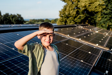 Cute boy on roof with solar panels, hand shielding eyes from summer sun. Rooftop solar or...
