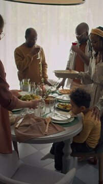 Full vertical slowmo of African American family sitting down to festive dinner at home, mother and daughter bringing trays with hot food, while father and granddad are tasting homemade wine by window