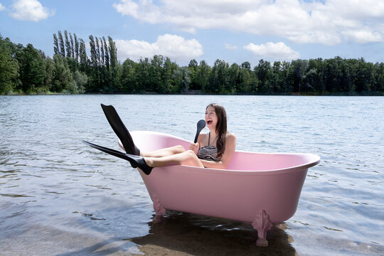 Cheerful girl with diving flipper holding back brush and sitting inside pink bathtub in lake