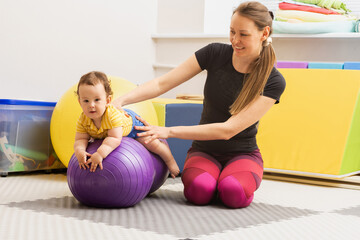 Happy child sitting on a violet fitness ball with his physiotherapist during a session of physical rehabilitation and exercise. Physical rehabilitation of children and early intervention