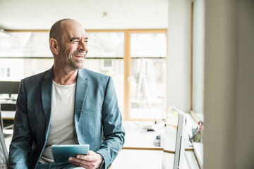 Happy mature businessman looking away sitting with tablet PC at office