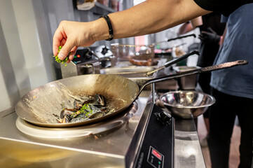 A hand sprinkling herbs into a frying pan with food on a stove, capturing the action of cooking