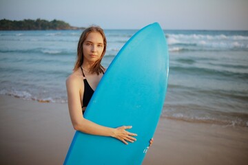 A young surfer woman stands on the tropical beach by the ocean with a blue surfboard before surfing.