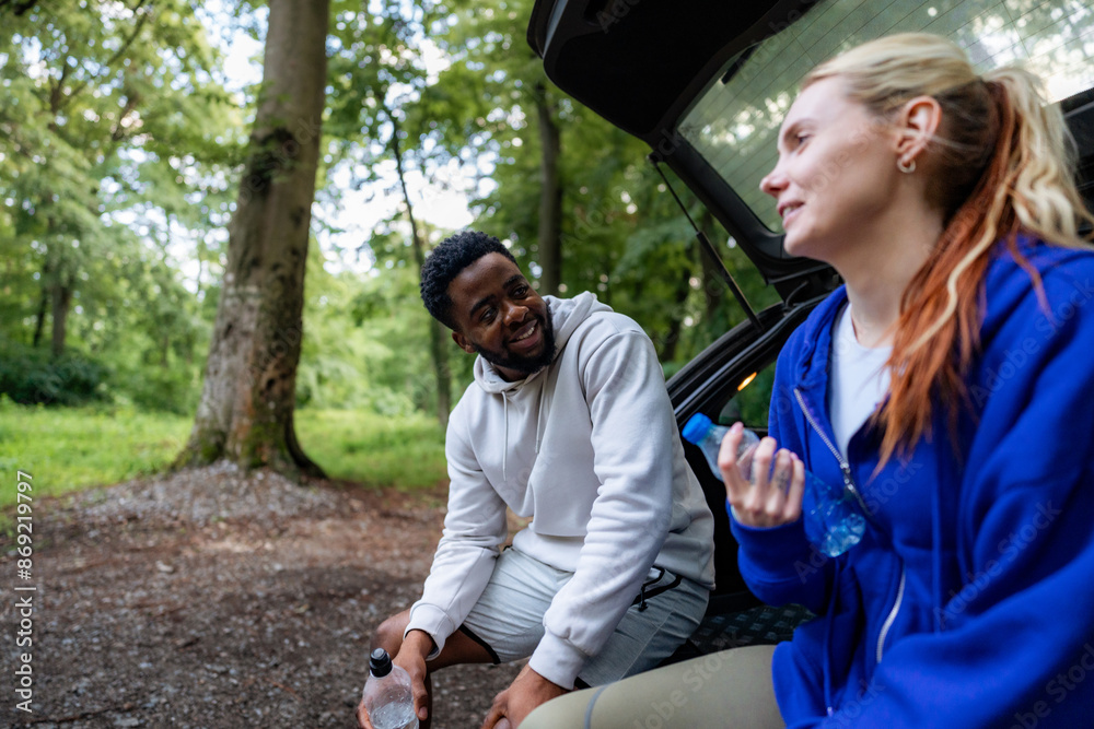 Wall mural Couple taking a water break after a run, sitting at the back of their car and chatting. They are dressed in sportswear and enjoying their outdoor workout.