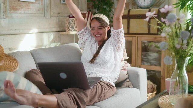 Cheerful woman looking forward to good news surprised excited looking at laptop, receives mail with positive results while sitting on couch in boho interior at home