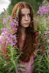 A tall, slender girl of 16-17 years old with long red curly hair in a pink shirt stands in the bushes of pink fireweed flowers, on a sunny summer day, half-length portrait, pink and green colors.
