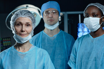 Waist up portrait of diverse medical team in operating room looking at camera with focus on mature woman surgeon in foreground, copy space