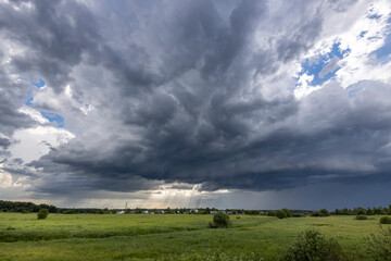 Fototapeta premium A wide shot of a dramatic spring sky, with large, dark clouds casting shadows over a lush green meadow. The clouds are full of movement, suggesting an impending storm.