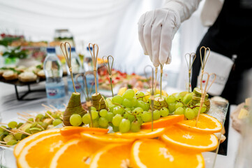A close-up of a person in white gloves arranging a catering display with skewers of green grapes and cheese on an orange slice base